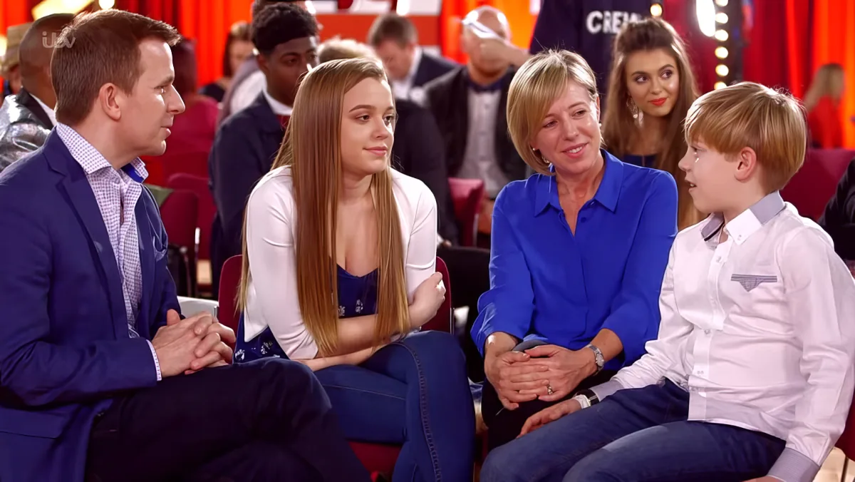 A family of four sits together on a stage, engaged in conversation, with a red curtain and audience in the background.