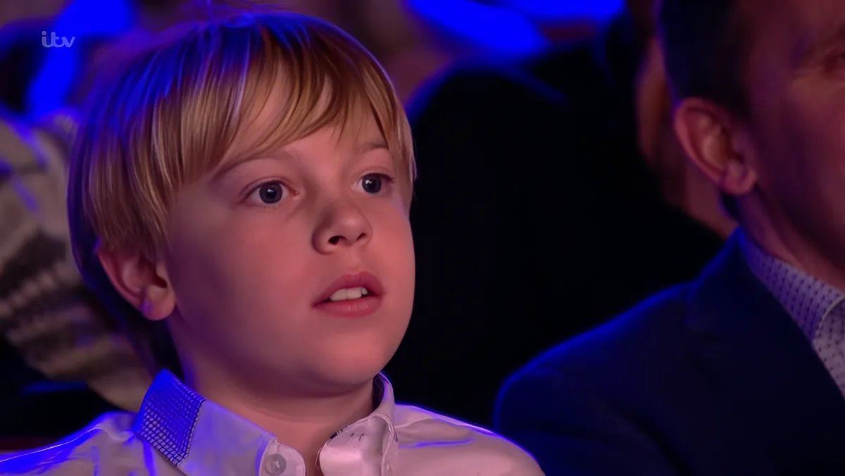 A young boy with blonde hair and a white shirt looks to the side, surrounded by blue lighting.