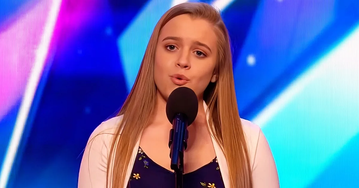A young woman with long blonde hair stands in front of a microphone, wearing a white cardigan over a floral dress...