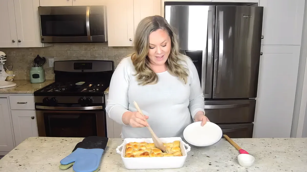 A woman in a kitchen prepares food, using a wooden spoon in a white dish on a granite countertop.