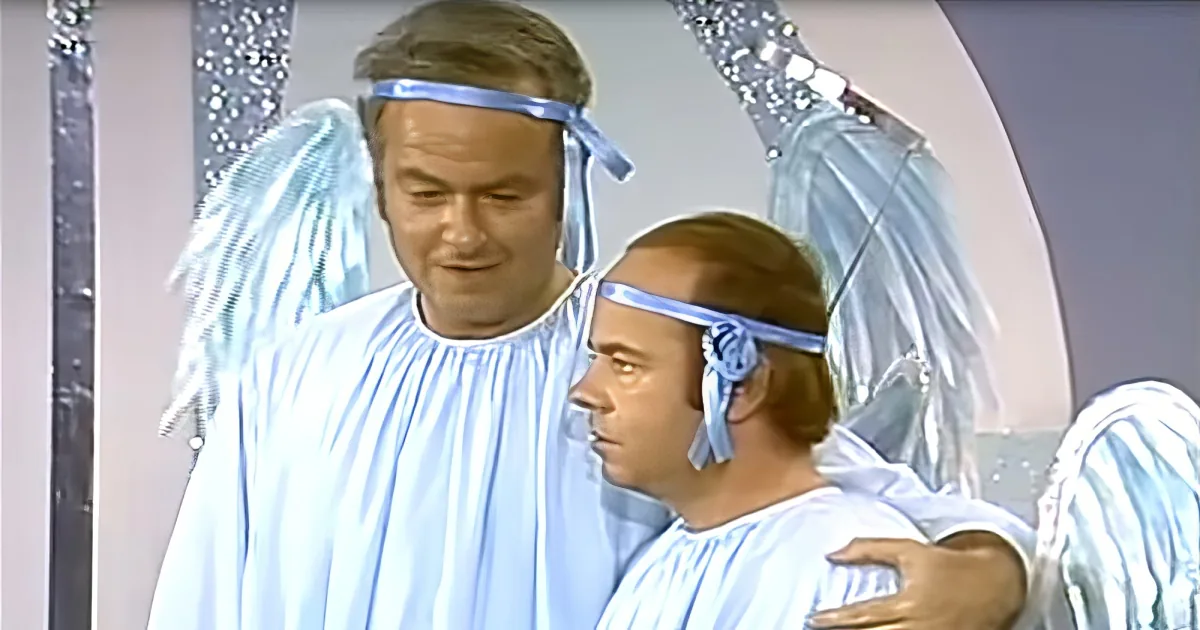 A man in a white angel costume with blue wings and a halo, smiling.