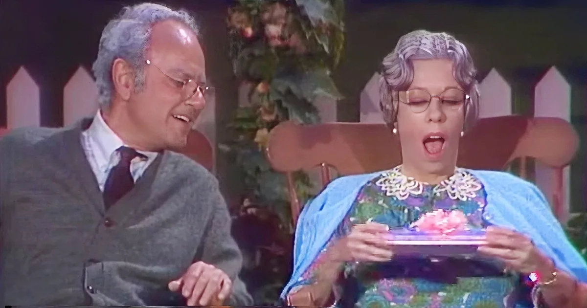 A man in a suit and a woman in a floral dress are sitting, with the woman holding a cake.