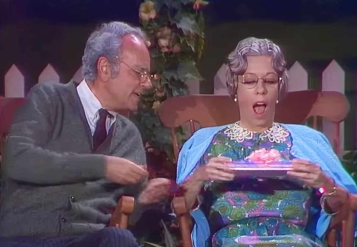 A man and woman are seated, the woman in a colorful dress holding a small object, both appearing surprised or excited.