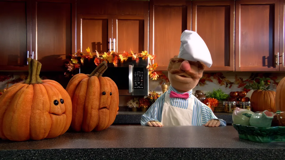 A chef puppet leans on a kitchen counter with pumpkins and autumn decorations in the background.