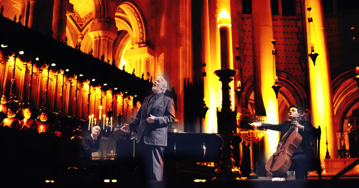 A man in a suit stands on stage, illuminated by warm lights, with a large organ and a cello player in the background.