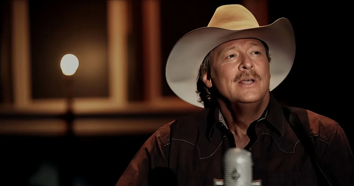 A man in a cowboy hat and brown jacket is speaking, with a warm, dimly lit background.