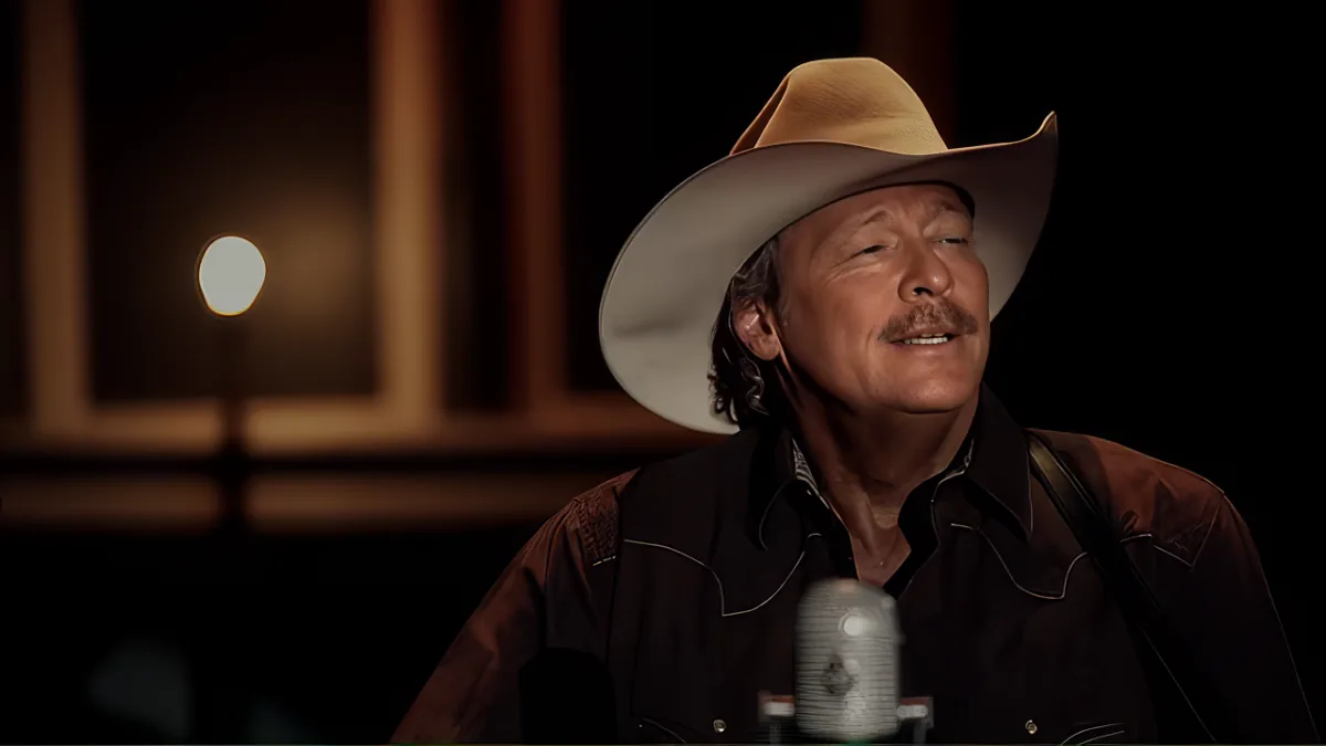 A man in a cowboy hat and brown shirt sings into a microphone, eyes closed, against a dark background with a light source.