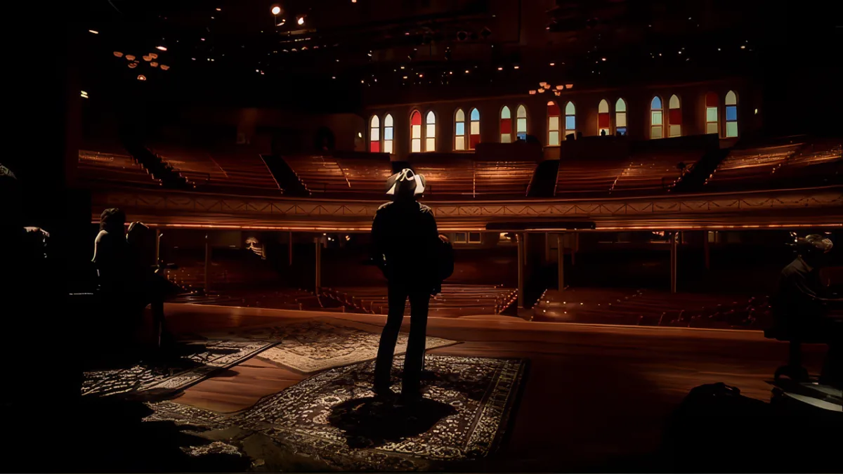 A person stands on stage in a dimly lit auditorium with stained glass windows in the background.