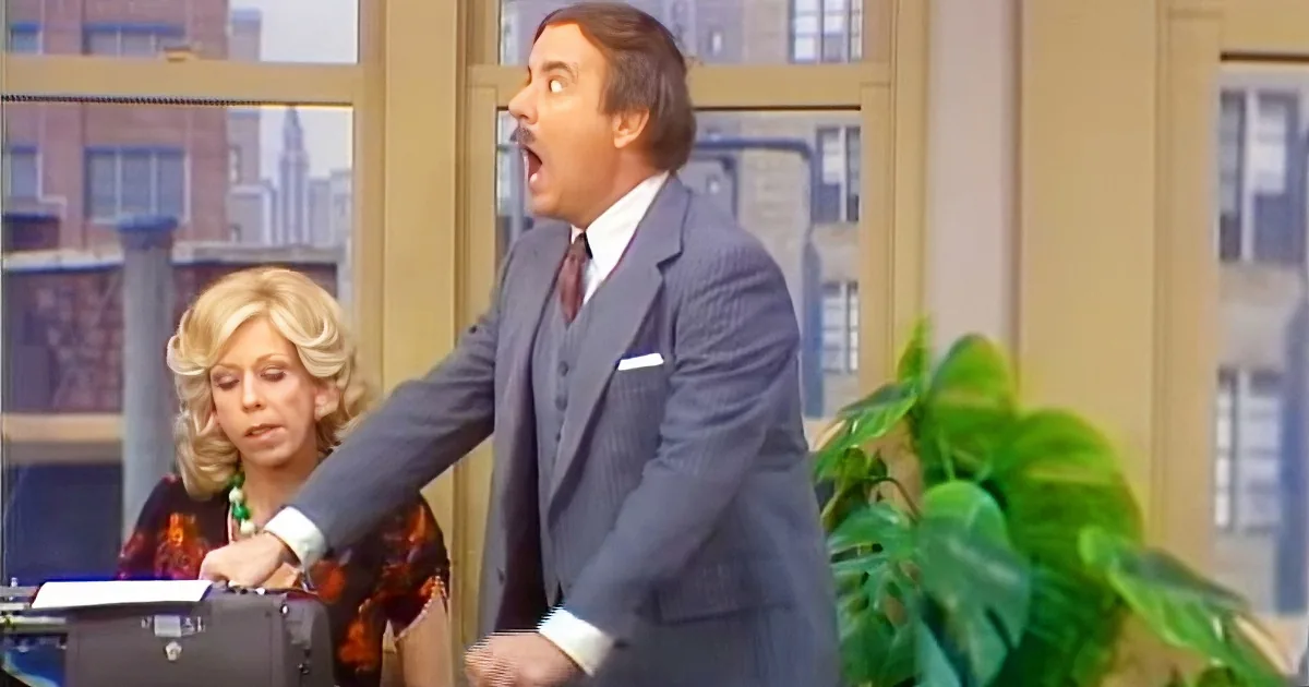 A man in a suit is leaning forward, mouth open, seemingly speaking or reacting, with a woman in the background.