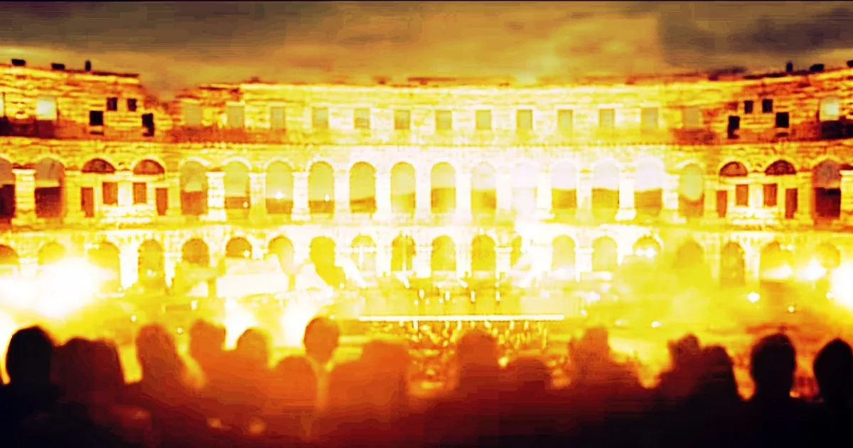 A large, ancient amphitheater with arched windows is illuminated by a bright, fiery glow, with silhouettes of people in...
