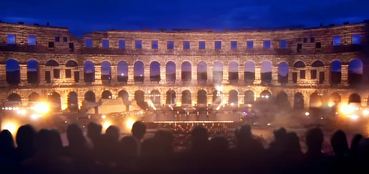 A large, illuminated amphitheater at night with silhouettes of people in the foreground.