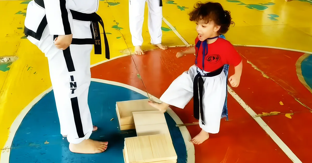 A child in a red and white uniform practices karate, stepping on wooden blocks with an adult in a white uniform nearby.