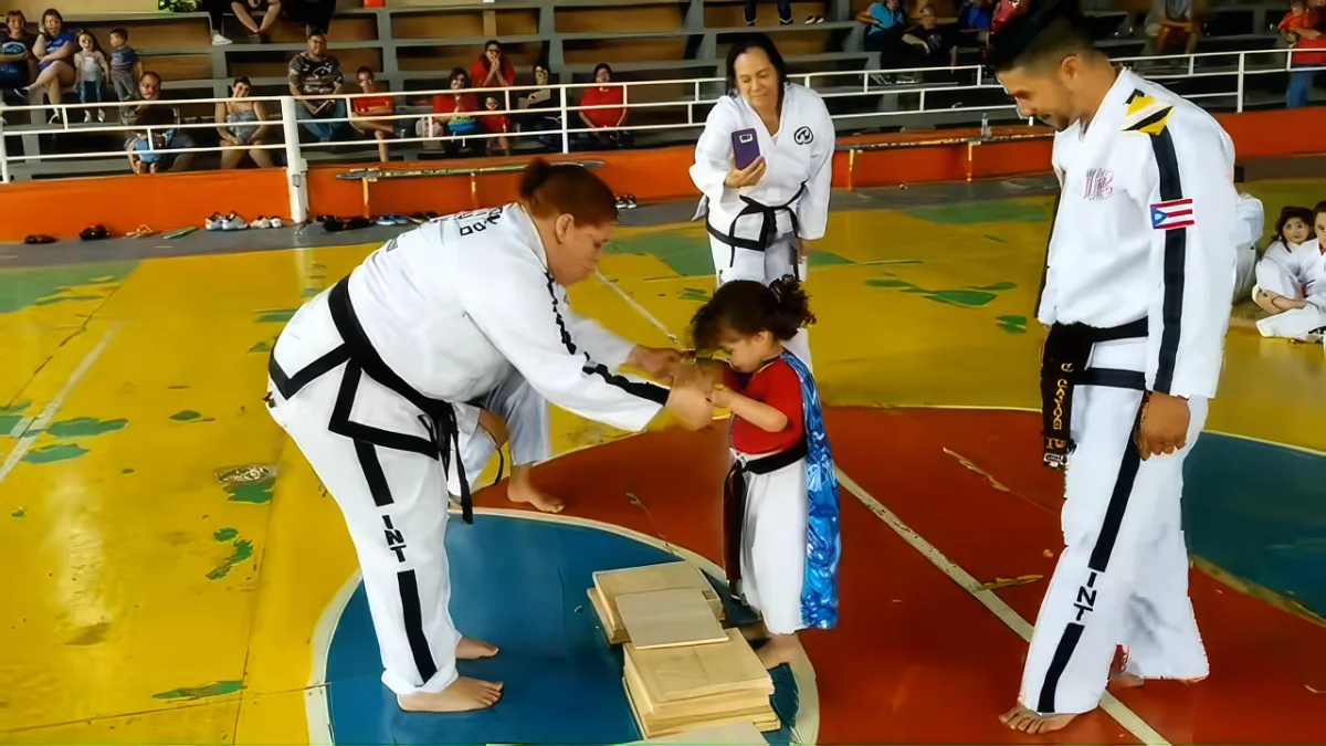 A young girl in a red shirt is being helped by a man in a white uniform to break a board during a martial arts demonstration.
