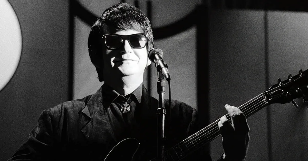 A man in sunglasses plays guitar, smiling, in front of a microphone.