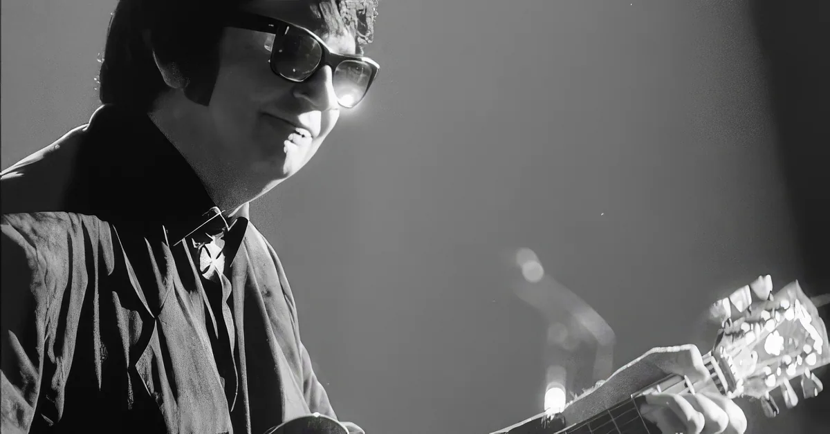 A man wearing glasses plays a guitar, smiling, in a black-and-white image.