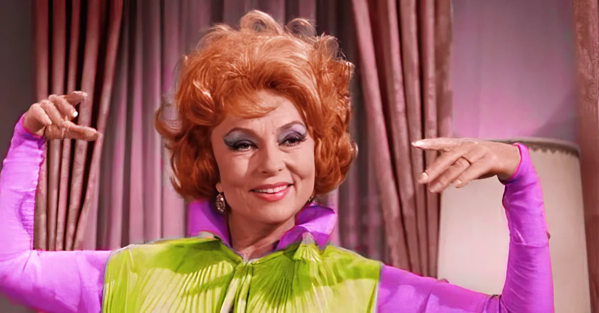 A woman with red hair and a green and purple outfit is posing dramatically in front of a curtain.