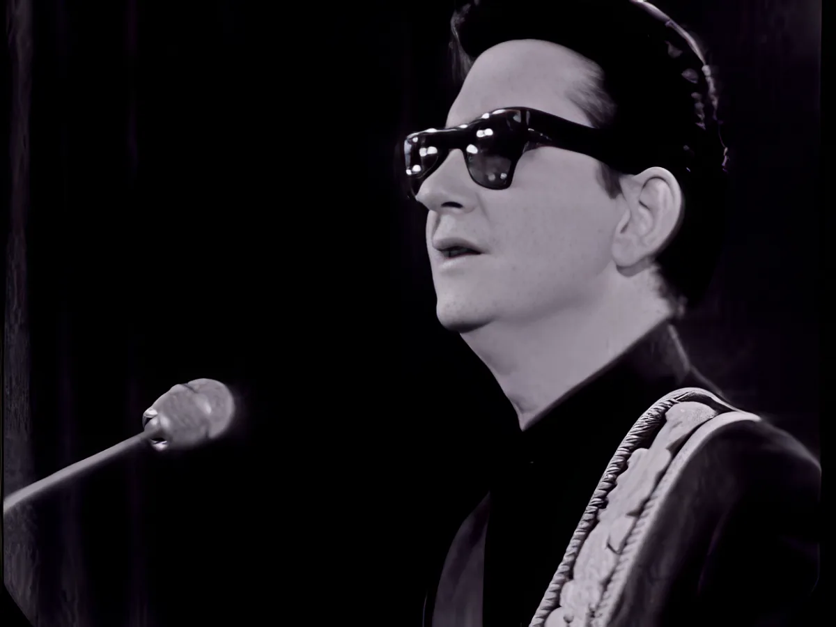 A man in sunglasses and a hat, wearing a black suit, is singing into a microphone.