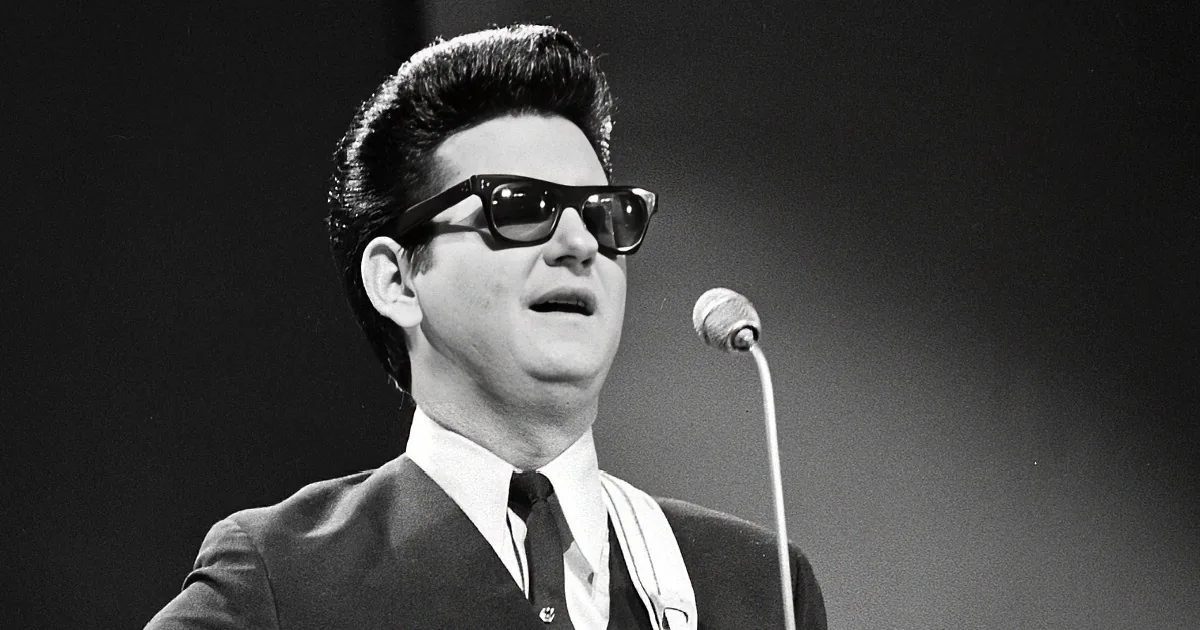 Roy Orbison, wearing sunglasses and a suit, sings into a microphone on stage.