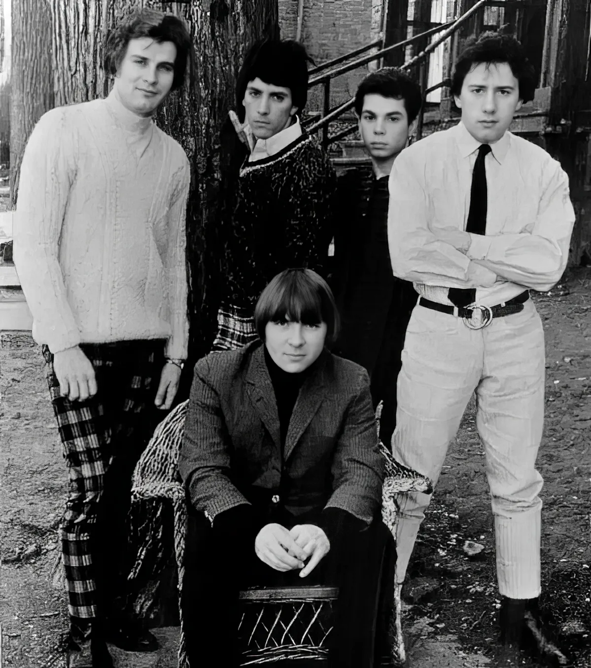 A group of five young men in casual attire, posing outdoors, with one sitting on a chair.