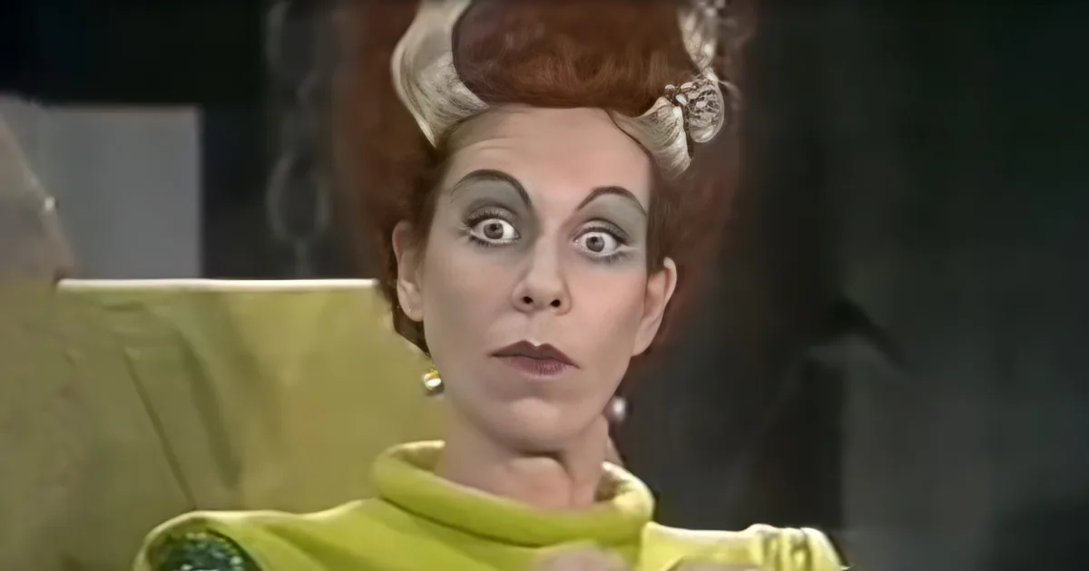 A person in a green outfit with exaggerated makeup and a large bow in their hair is looking surprised.