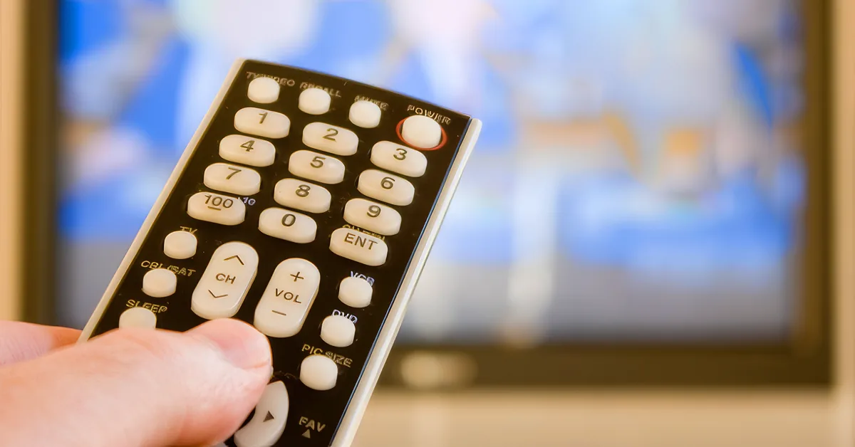 A hand holds a black TV remote control in front of a blurred TV screen.