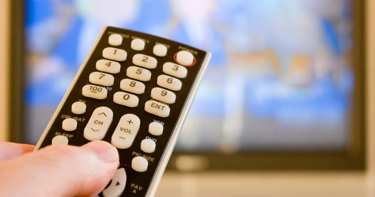 A hand holds a black TV remote control in front of a blurred TV screen.