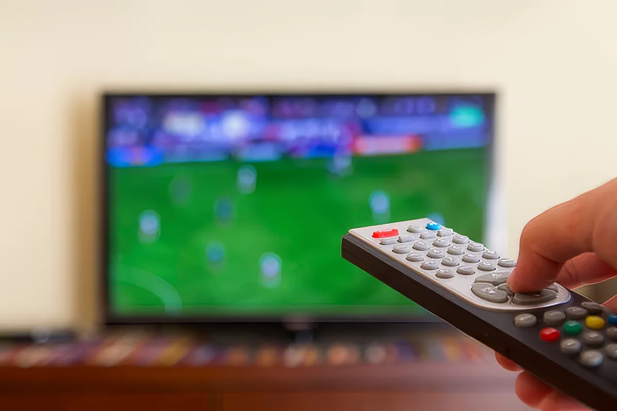 A hand holds a remote control in front of a blurred TV screen displaying a soccer game.