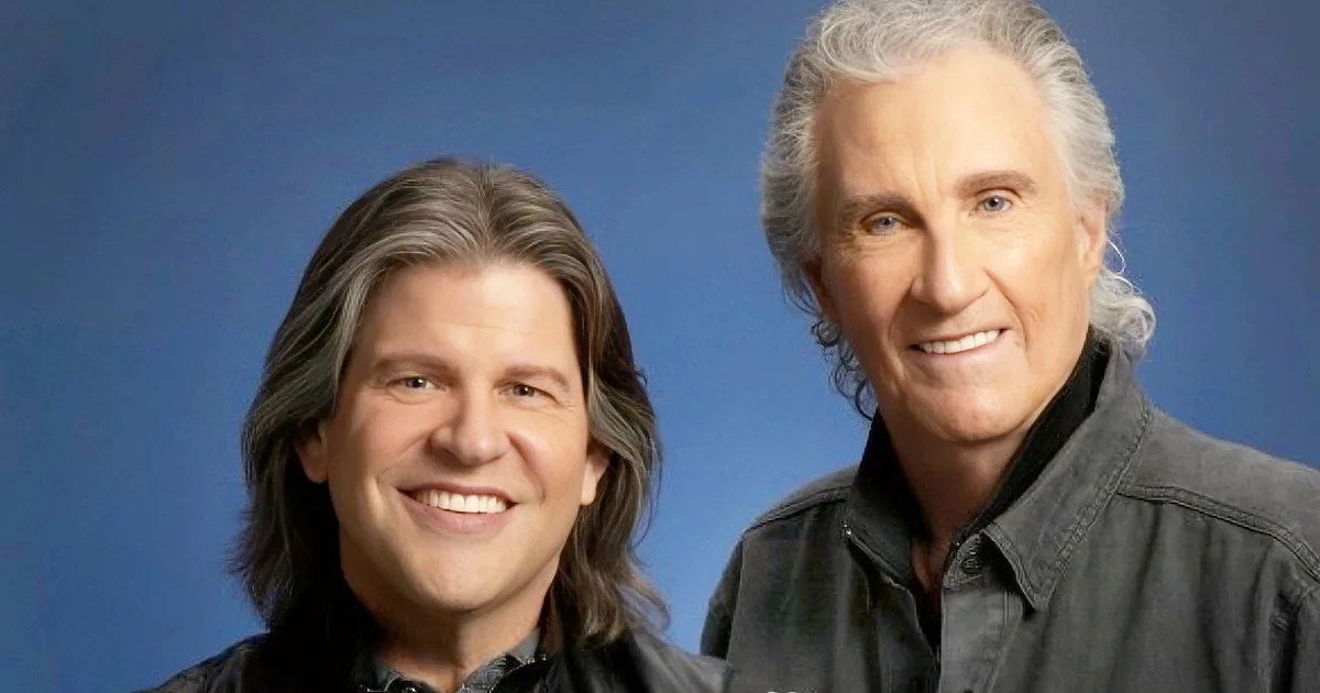 Two men with long hair, smiling, wearing dark jackets, against a blue background.