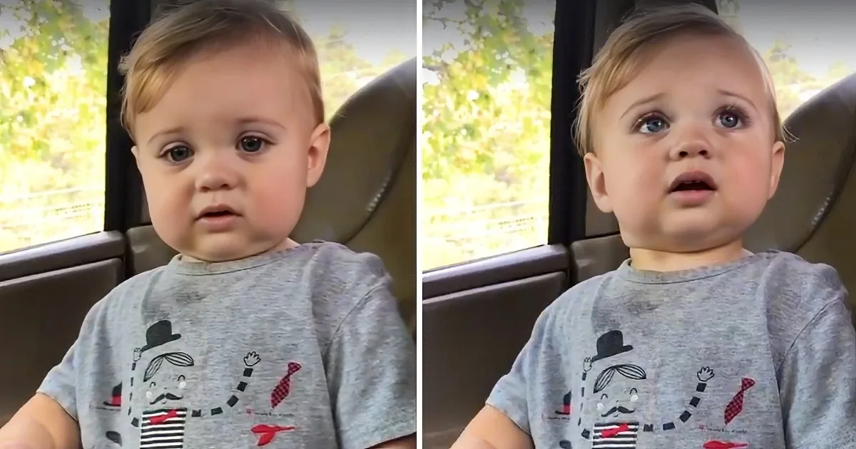 A baby in a grey shirt with a cartoon character sits in a car seat, looking slightly to the side.