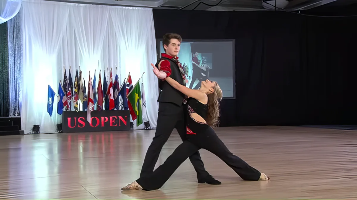 A couple in formal attire dances gracefully on a stage with flags and a "JUS OPEN" sign in the background.