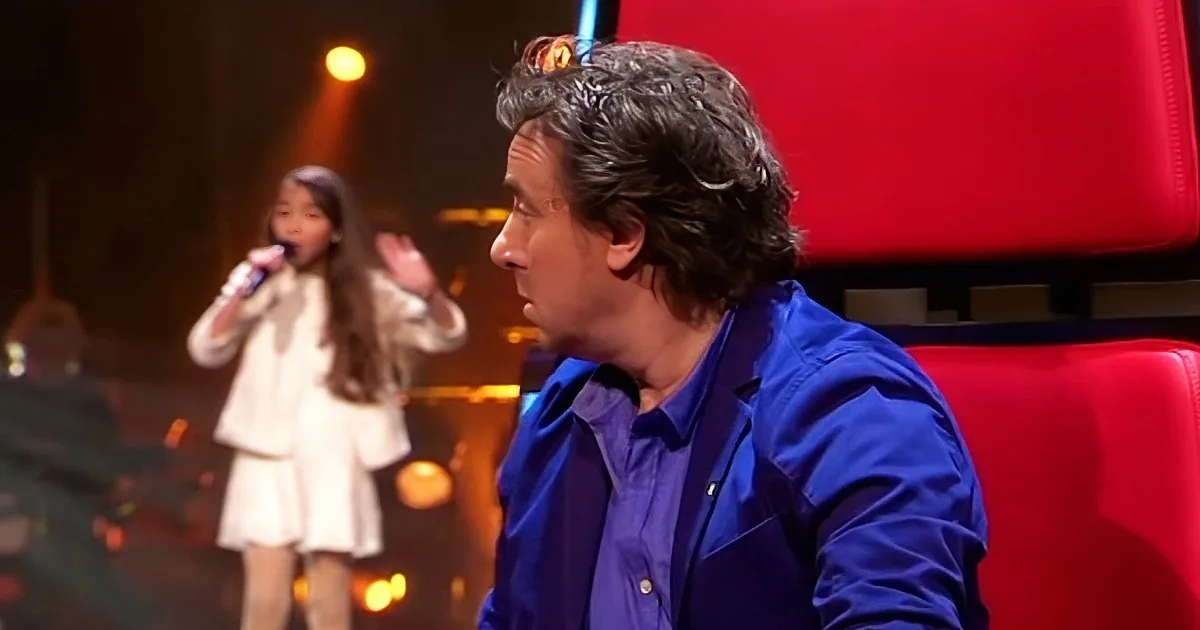 A man in a blue suit sits on a red chair, watching a young girl sing on stage.