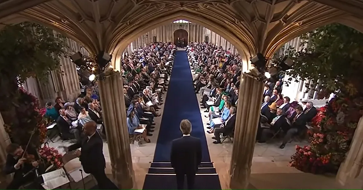 A man in a suit walks down a blue carpet in a grand hall with an audience on both sides.