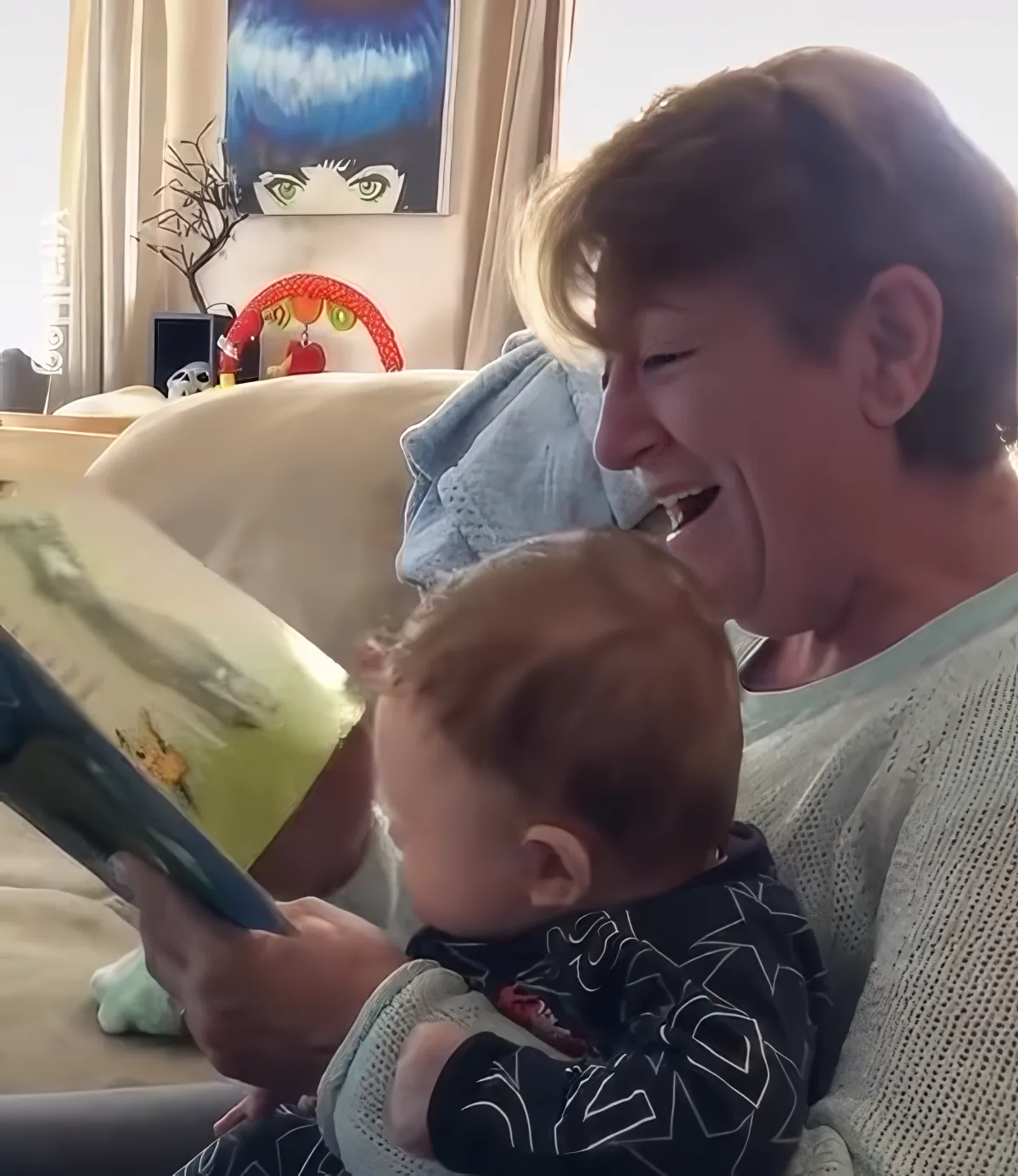 A woman is reading a book to a baby, both smiling, with a colorful background.