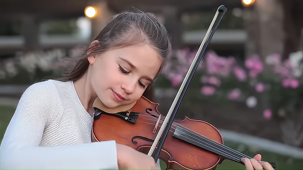 A young girl with long hair, wearing a white top, is playing a violin in an outdoor setting with blurred flowers in the...