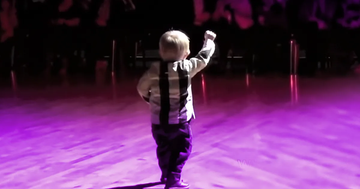 A toddler in a striped shirt and suspenders stands on a stage with a purple light, raising one arm.