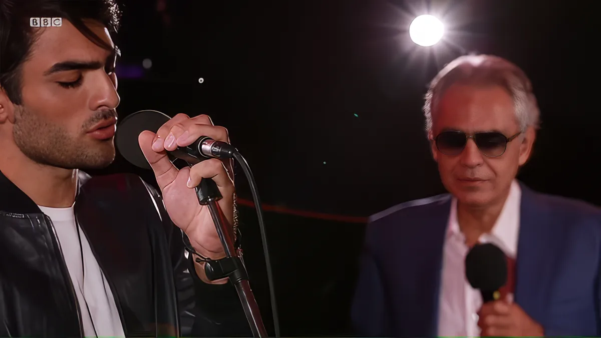 A man in a black leather jacket sings into a microphone, with another man in sunglasses observing in the background.