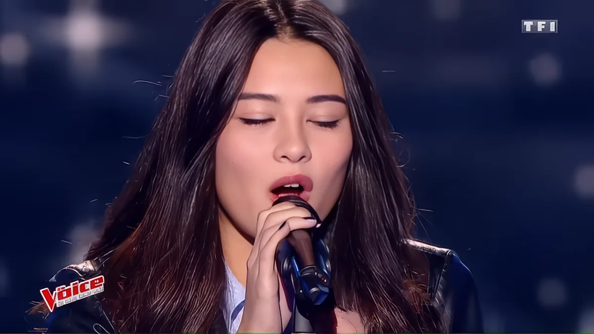 A person with long dark hair sings into a microphone, eyes closed, in a dimly lit setting.