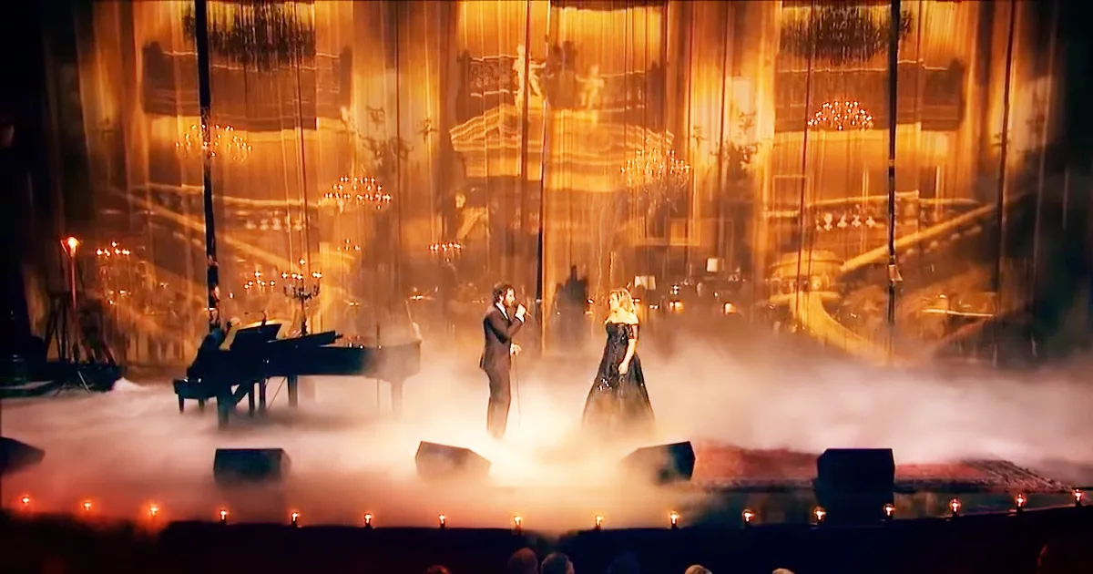 A man and woman sing on a smoky stage with a grand piano and a backdrop of golden lights.