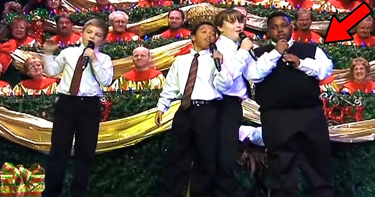 A young boy in a black vest and white shirt sings into a microphone, surrounded by a choir in festive attire.