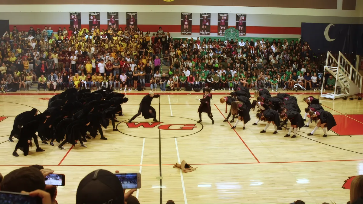 A group of dancers in black costumes perform a synchronized routine on a gymnasium floor.