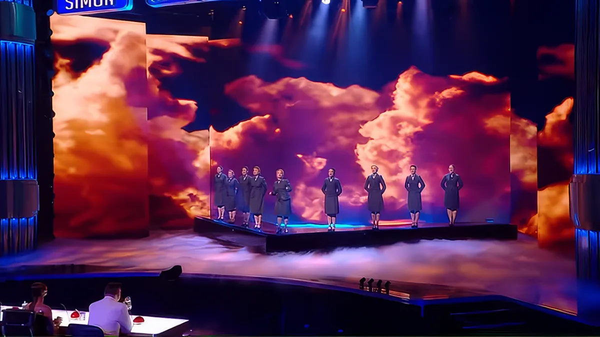 A group of performers in black robes stand on a stage with a dramatic sky backdrop and clouds.