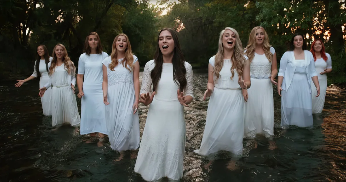 A group of women in white dresses sing joyfully in a shallow stream surrounded by trees.