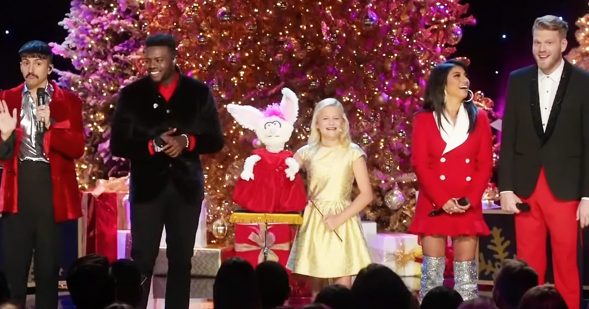 A young girl in a gold dress stands on stage with a Christmas tree backdrop, smiling.