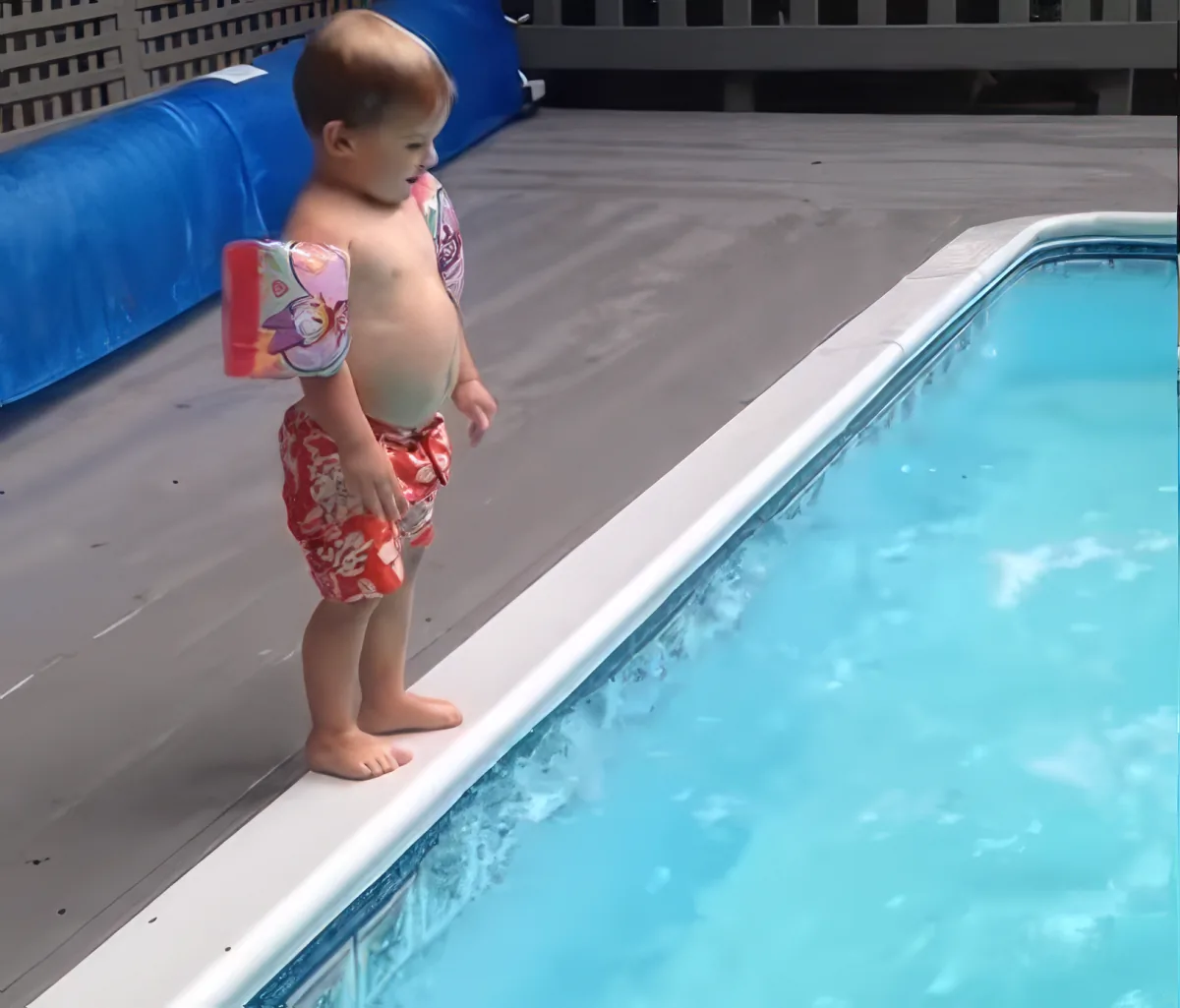 A toddler in swimwear stands at the edge of a pool, holding a floatation device.