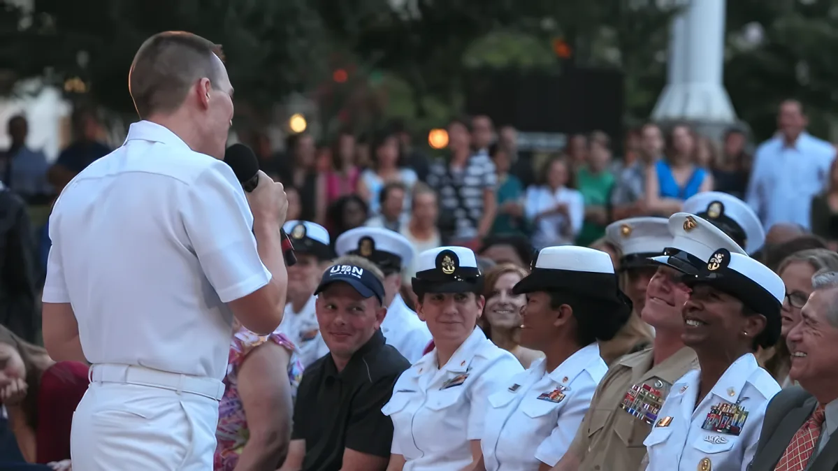 A man in a white uniform speaks into a microphone to a group of uniformed sailors and a crowd of onlookers.
