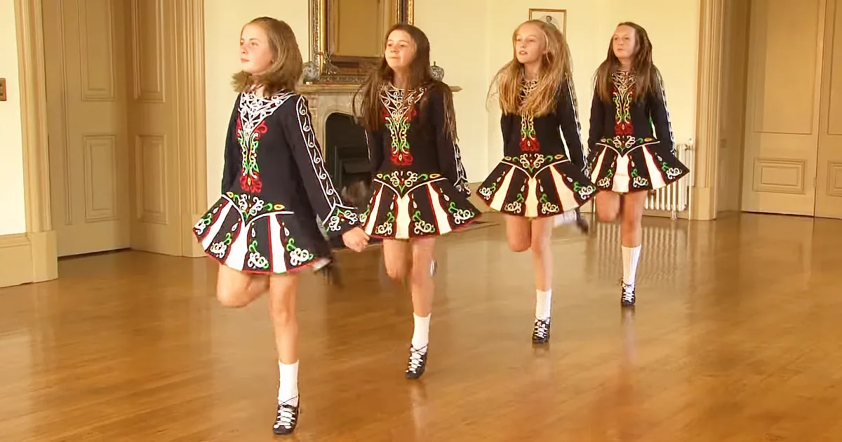 Four young dancers in traditional Irish attire perform a synchronized step in a spacious room with wooden floors and...