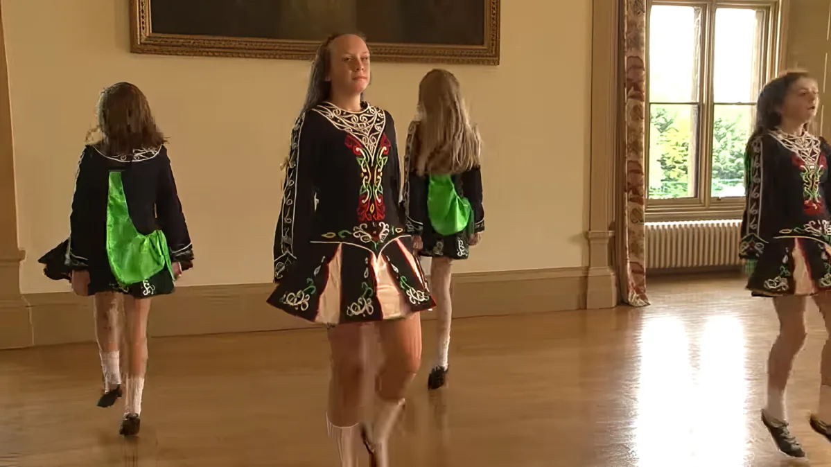 A girl in a traditional Irish dance costume is performing a step in a dance studio.