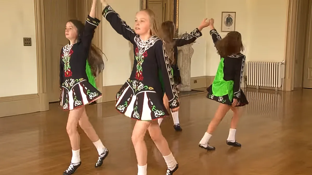 A young girl in traditional Irish dance attire performs a step in a studio setting.