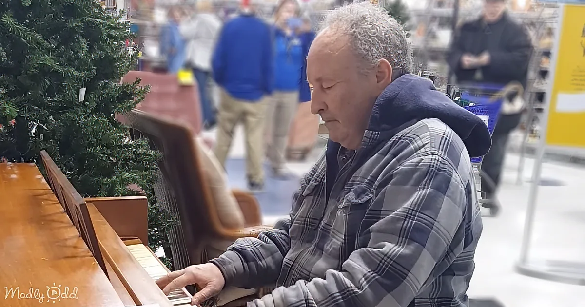 An elderly man in a plaid shirt plays a piano in a store with Christmas trees and people in the background.