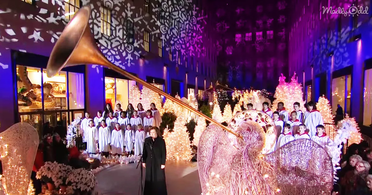 A choir in white robes sings under a giant golden trumpet, surrounded by Christmas lights and snowflake projections.